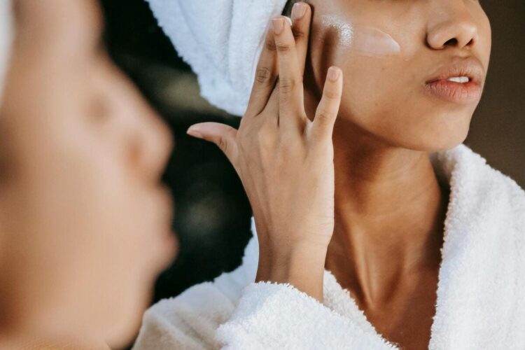 Crop ethnic woman applying cream on face against mirror